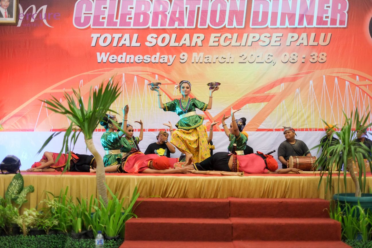 Dancers in green and gold hold brass bowls above a stage at the eclipse celebration dinner
