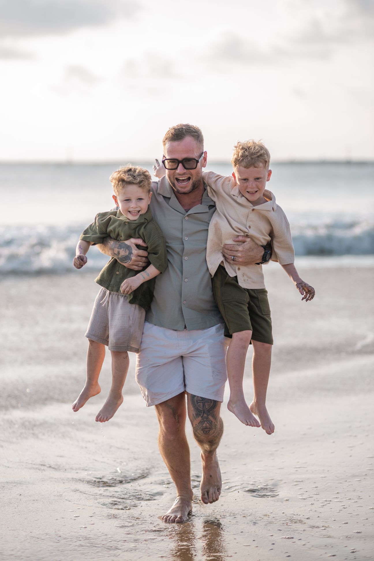 A father carries two young sons along the water’s edge at low tide