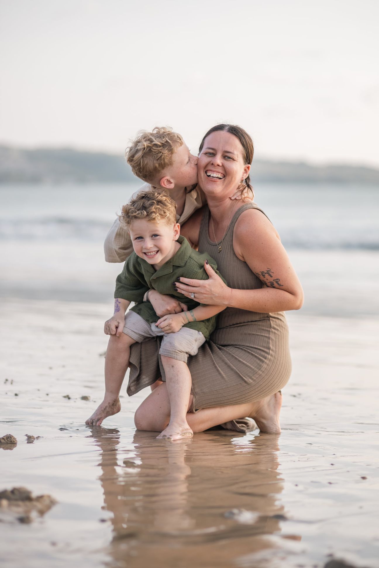 A parent carries two young children through shallow surf