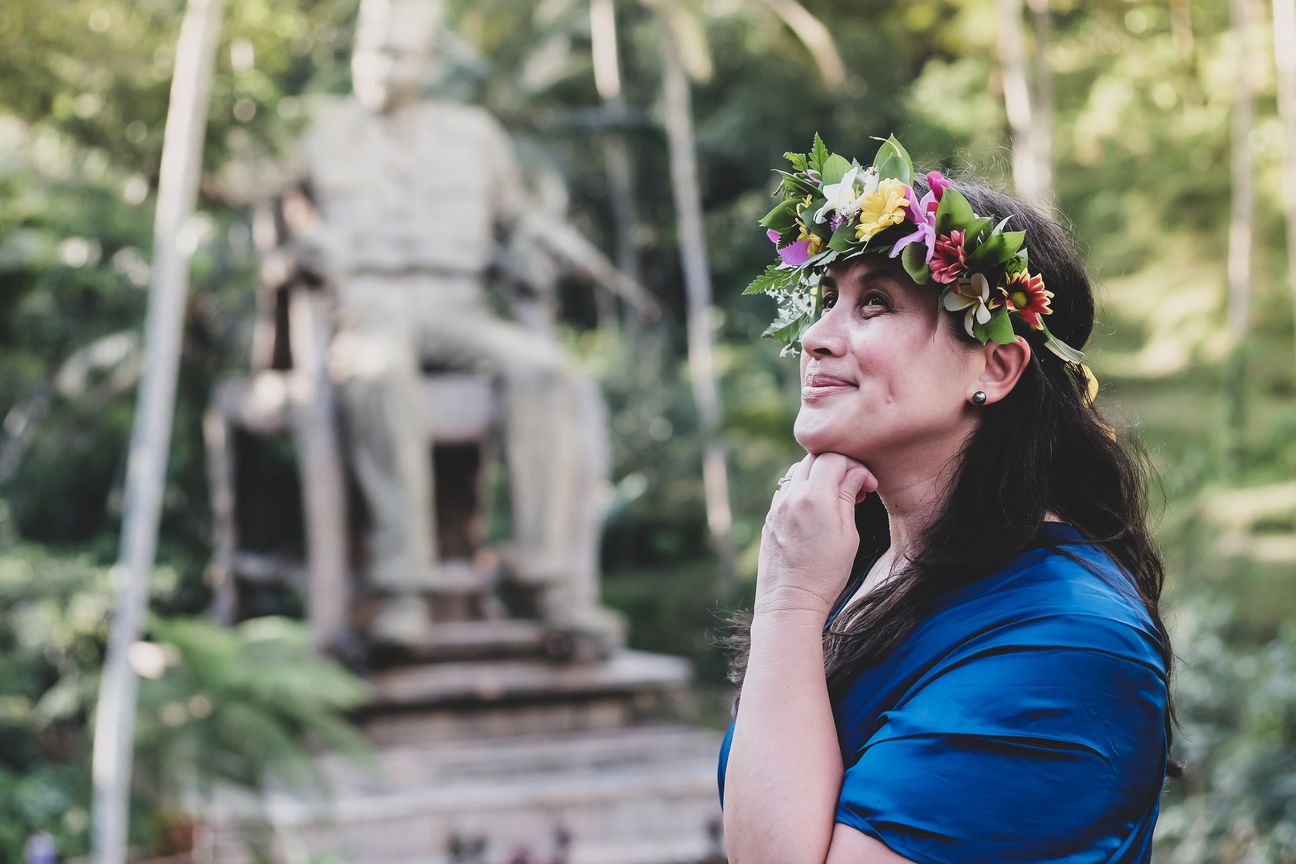 A woman in a flower crown looks up beside a stone statue in a garden