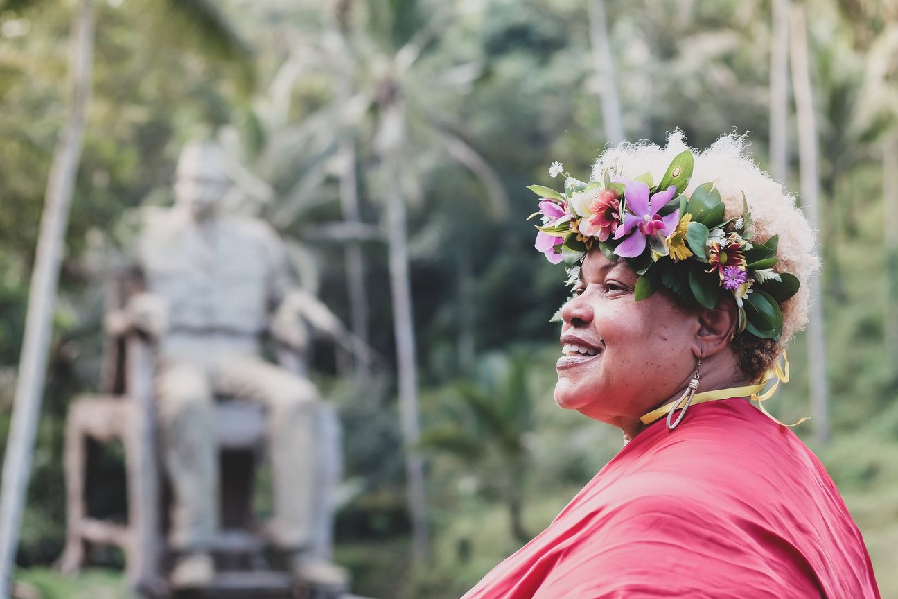 A woman in a flower crown looks up beside a stone statue in a garden