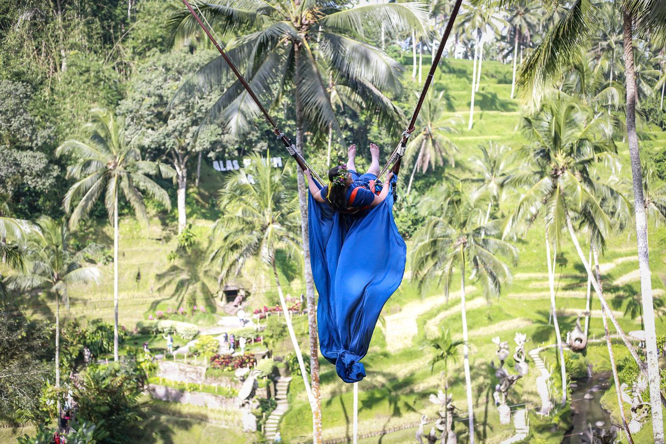 A woman in a long flowing dress swings out above palm trees and rice terraces