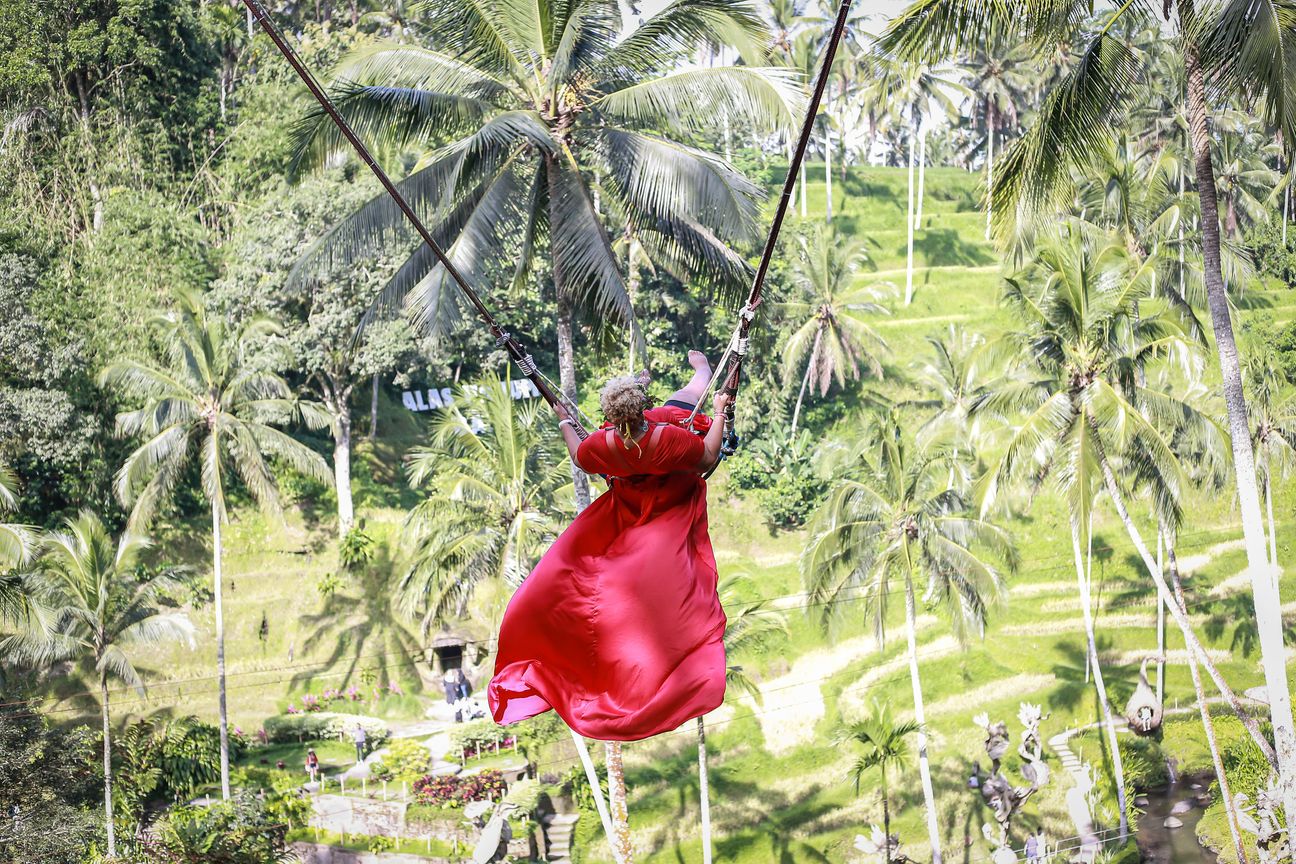 A woman in a long flowing dress swings out above palm trees and rice terraces