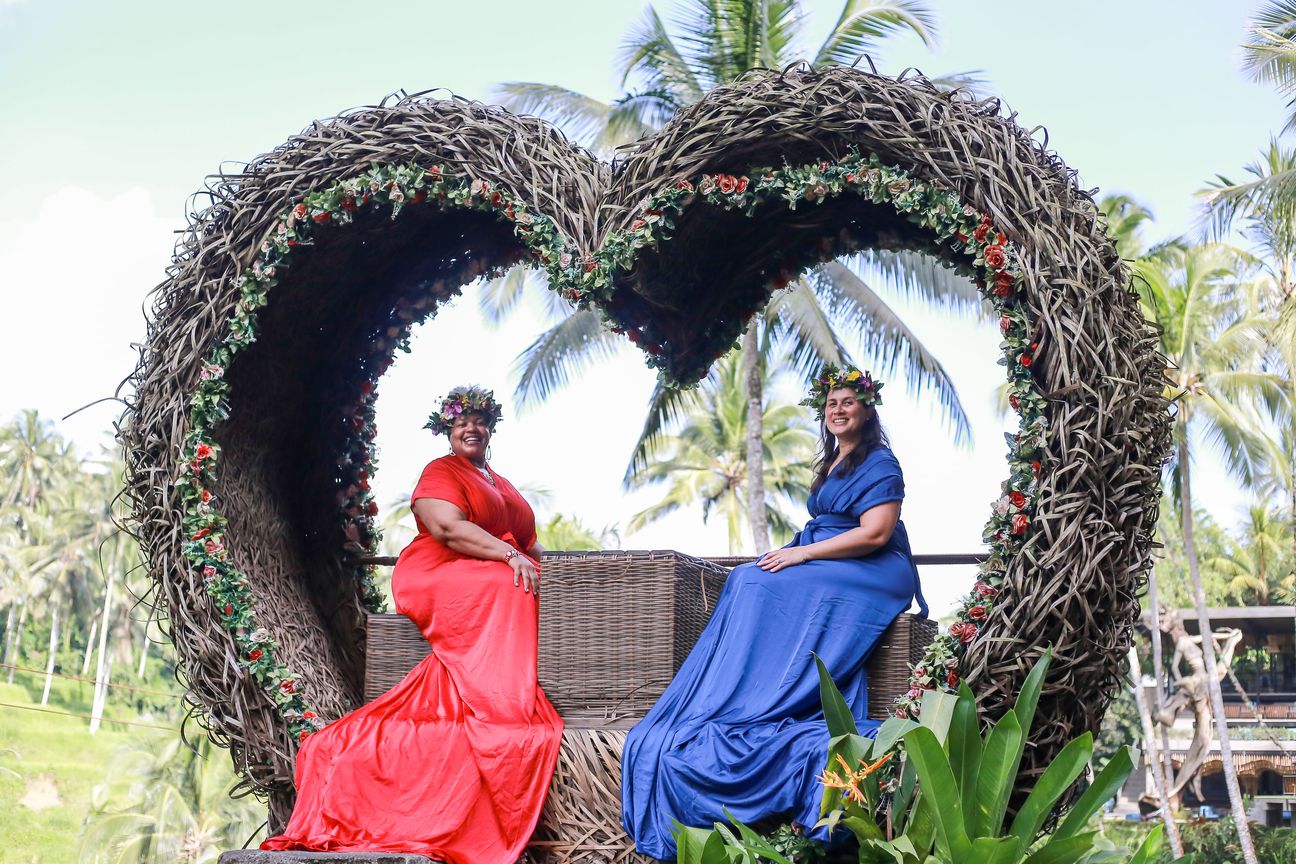 Two women in long red and blue gowns pose together in a woven photo frame