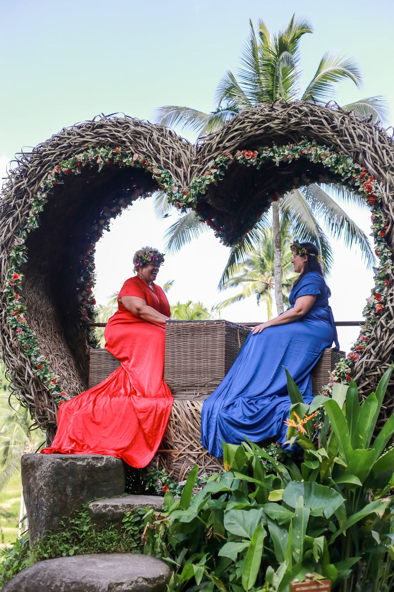 Two women in long red and blue gowns pose together in a woven photo frame