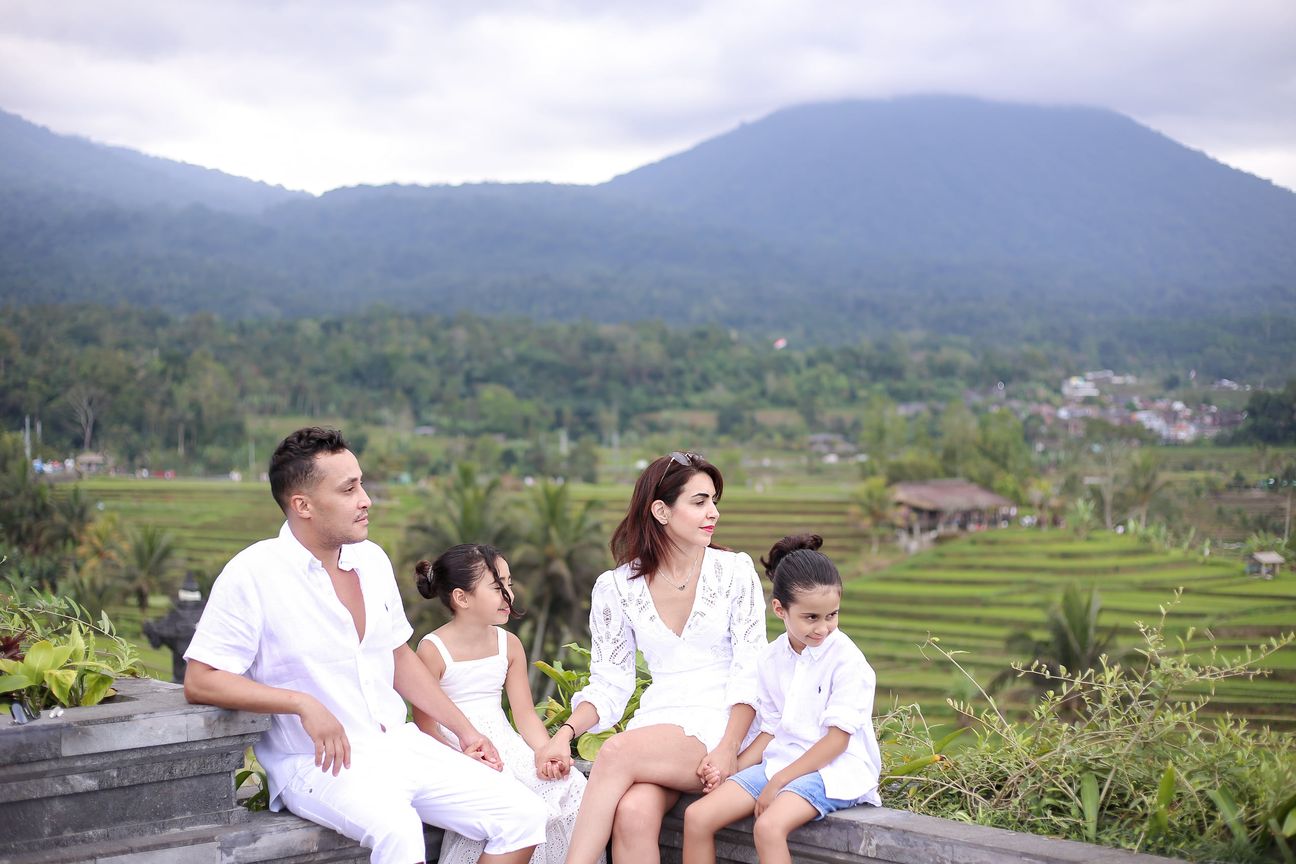 A family of four sit together on a stone terrace with hills and rice paddies behind them