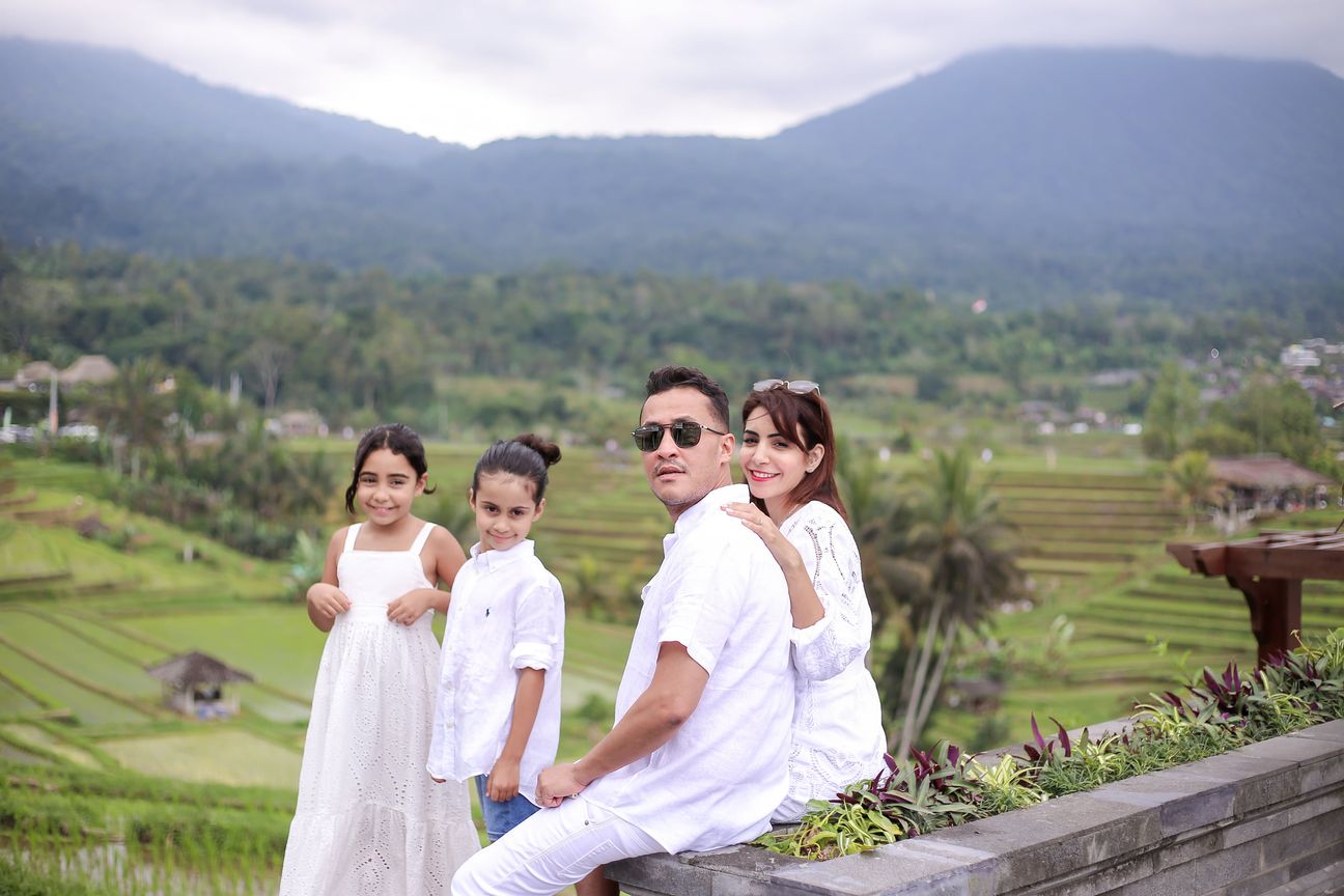 A family of four sit together on a stone terrace with hills and rice paddies behind them