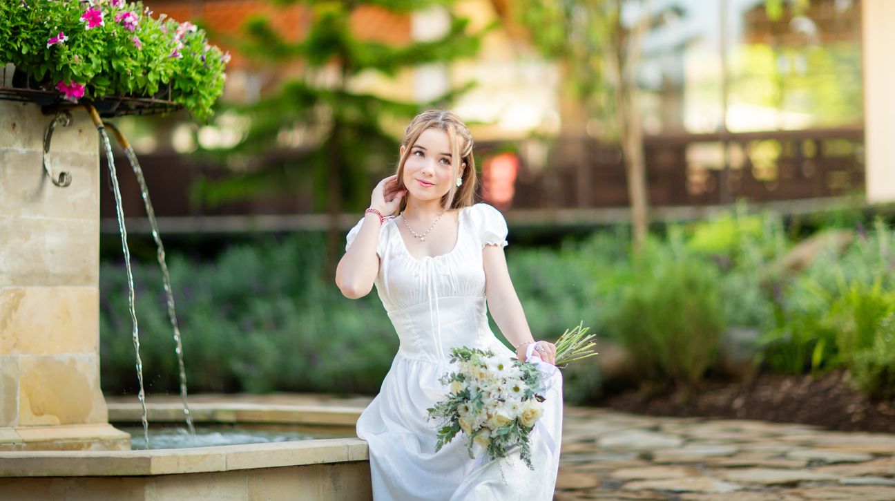 A woman in a white dress poses beside sunlit garden architecture