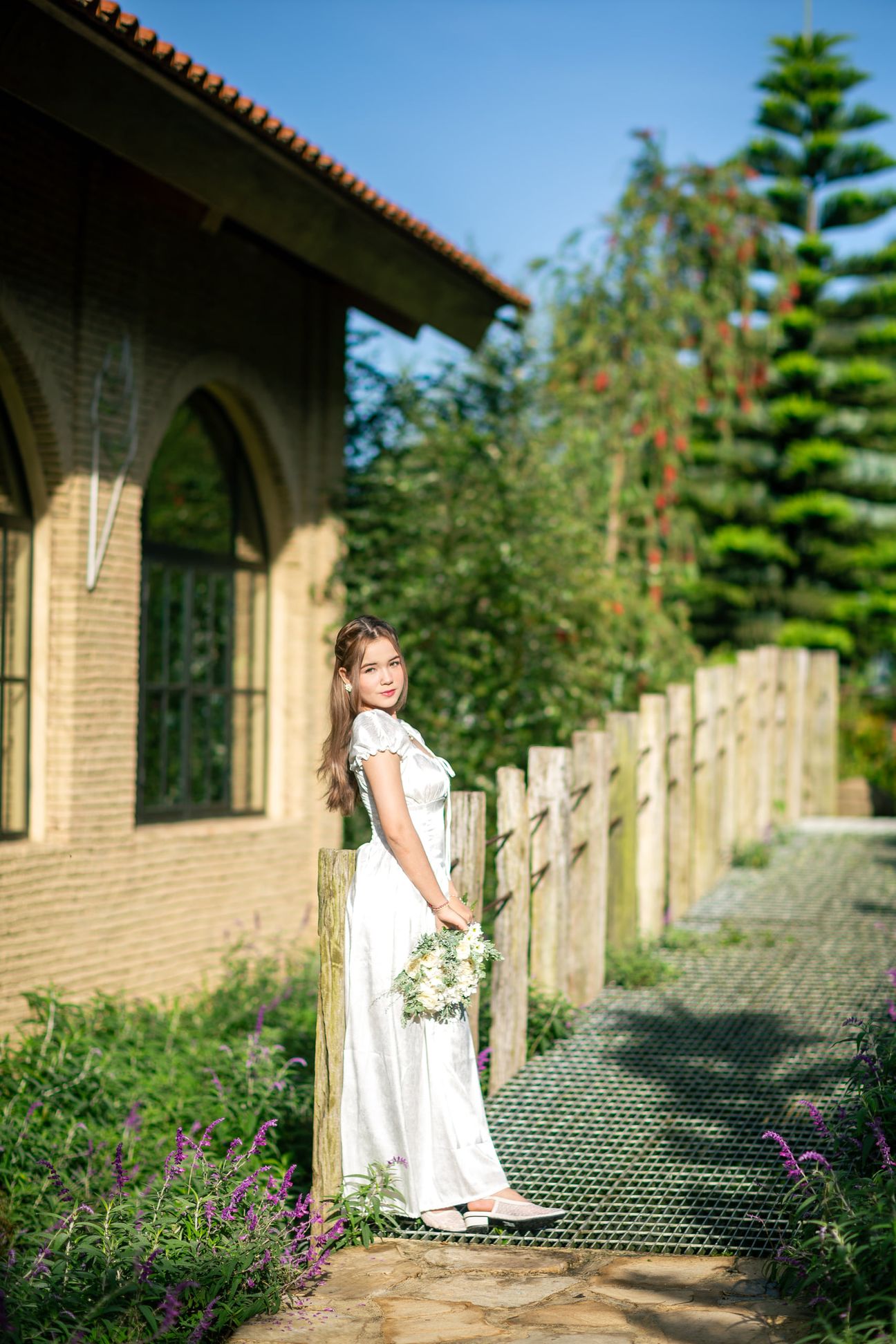 A woman in a white dress poses beside sunlit garden architecture