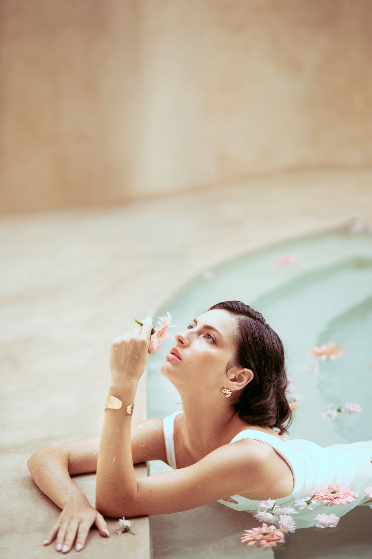 A woman in white holds a pink flower beside a pool scattered with petals