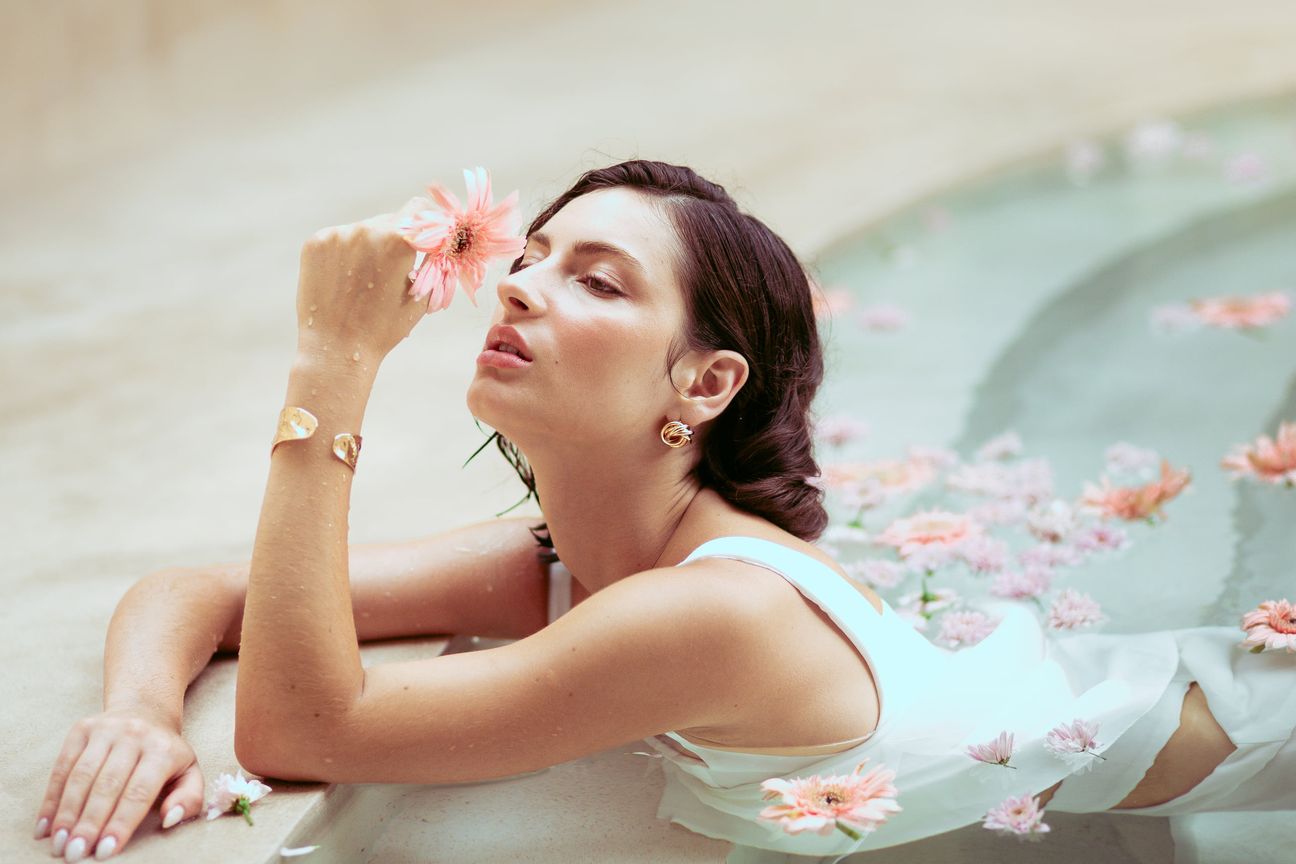 A woman in white holds a pink flower beside a pool scattered with petals
