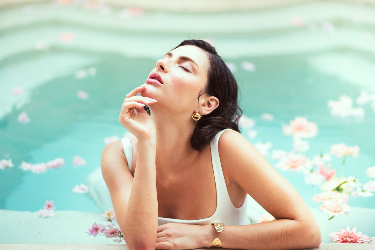 A woman in white holds a pink flower beside a pool scattered with petals