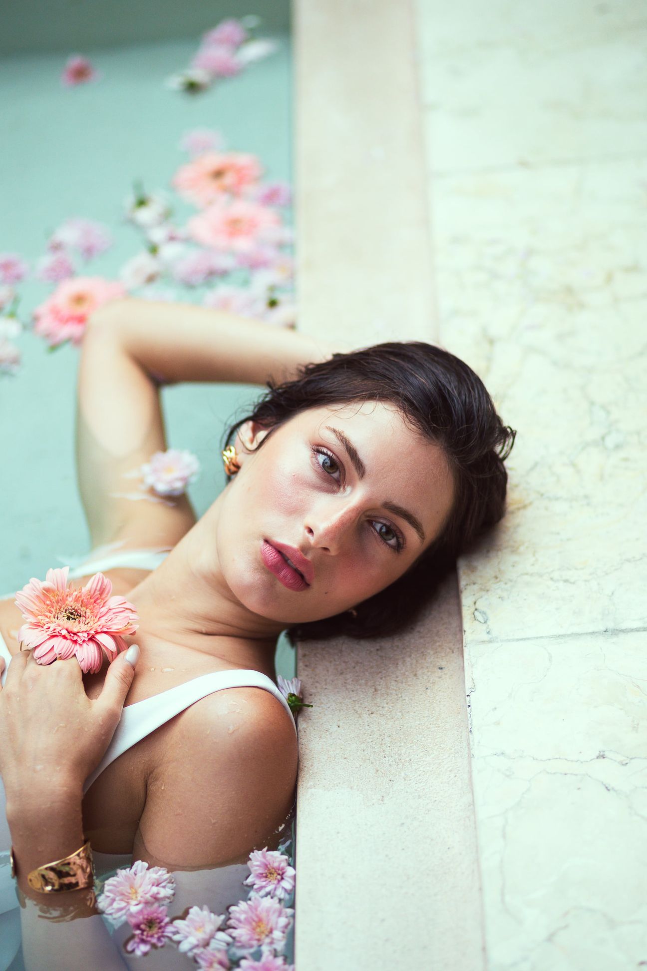 A woman in white holds a pink flower beside a pool scattered with petals