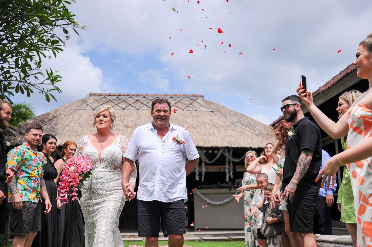 Newlyweds walk hand in hand beneath falling red petals, guests photographing them