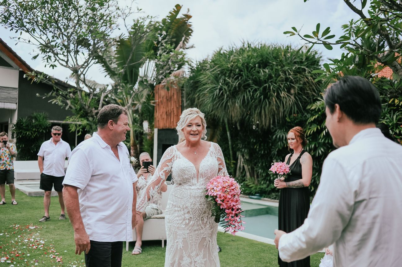 A bride carrying a bouquet walks through a garden as guests turn to watch